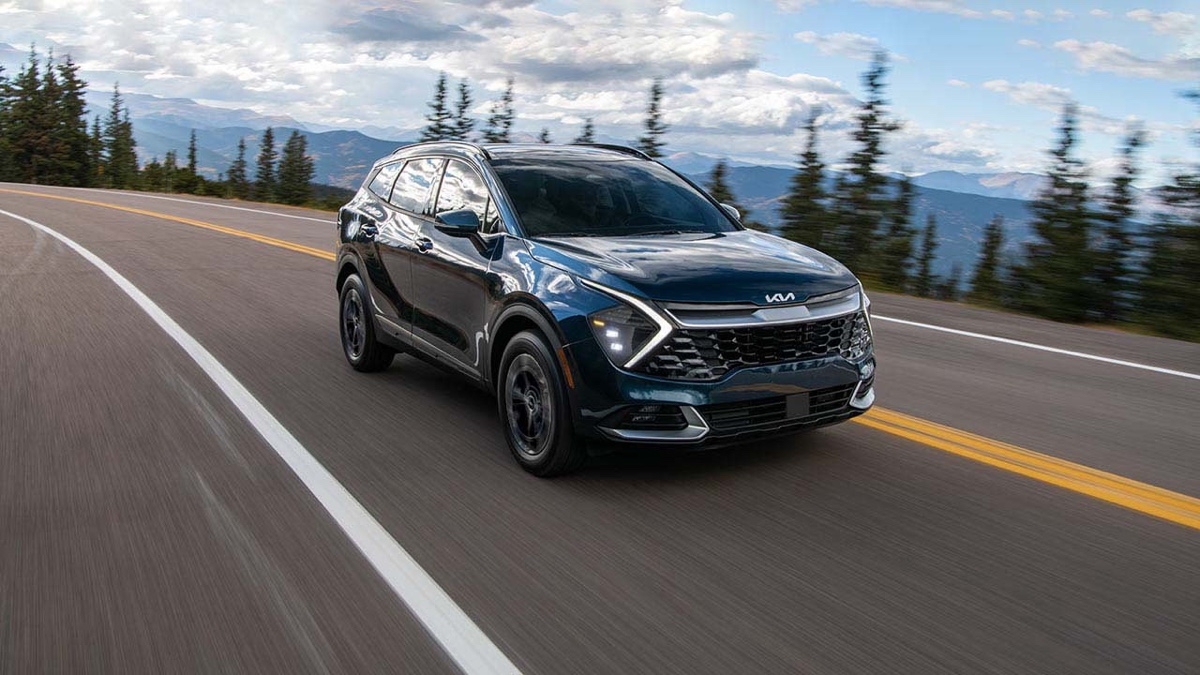 Kia Sportage hybride noir roulant sur une route montagneuse bordée d’arbres, avec un ciel nuageux en arrière-plan.