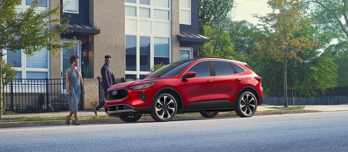 Couple regardant un Ford Escape hybride rouge stationné devant une maison urbaine, symbolisant l’accessibilité locale pour les essais routiers.