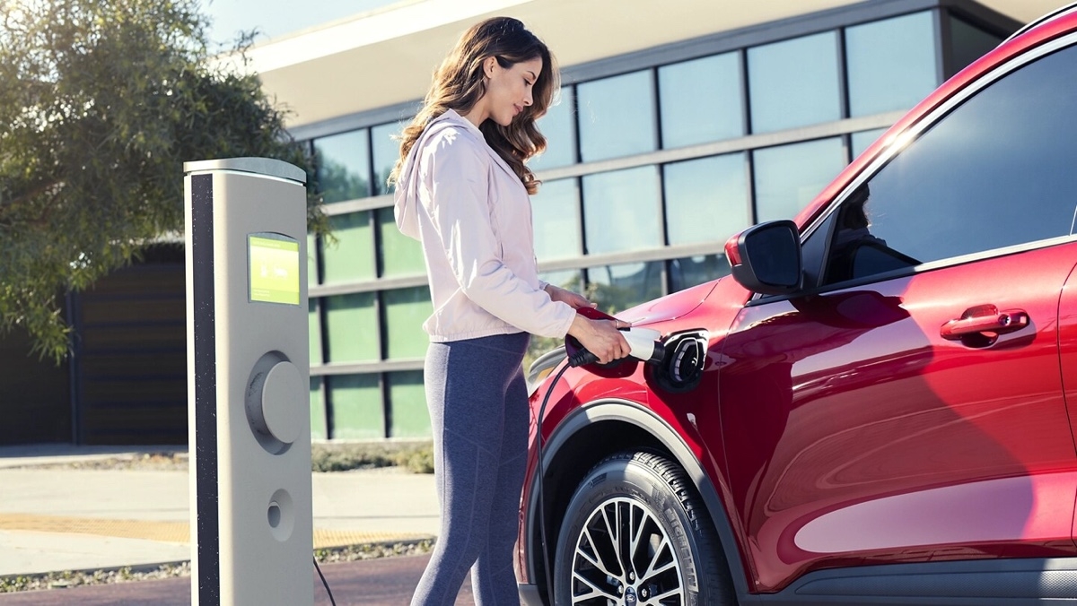 Femme branchant un VUS hybride rouge à une borne de recharge devant un bâtiment moderne, illustrant une étape d’achat réfléchi pour un véhicule électrifié.